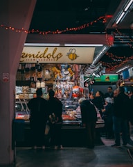A bustling market scene with a group of people gathered around a stall selling a variety of colorful candies and goods. The stall is decorated with festive red string lights, and the name of the shop is displayed prominently with a bee logo. The background is filled with more stalls and bright lighting, indicating an indoor setting.