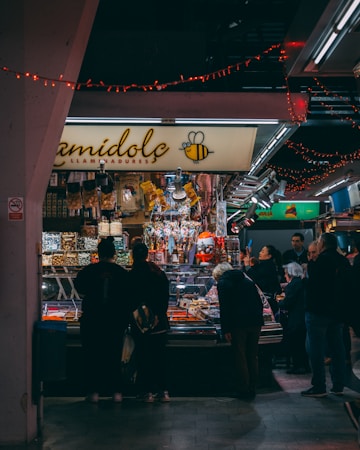 A bustling market scene with a group of people gathered around a stall selling a variety of colorful candies and goods. The stall is decorated with festive red string lights, and the name of the shop is displayed prominently with a bee logo. The background is filled with more stalls and bright lighting, indicating an indoor setting.