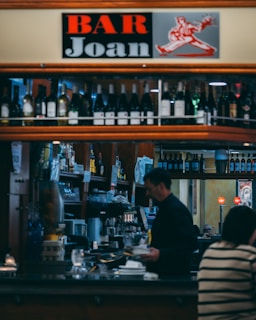 Interior view of a bar with a sign that reads 'Bar Joan' situated above a shelf filled with bottles of wine and liquor. There are two people in the image; one of them is a bartender, possibly pouring a drink or preparing something at the counter, while the other is sitting at the bar. The bar area is well-stocked with various bottles, glasses, and equipment.