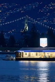 A well-lit Chevron gas station floats on a calm body of water, with city lights twinkling in the background. The structure is illuminated, casting reflections on the water's surface. Behind it, a large suspension bridge with lights spans across, accompanied by a backdrop of hills dotted with small glowing lights from buildings.