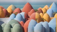 A collection of uniquely shaped, colorful dome-like structures, resembling a cluster of vibrant eggs or pods. Each structure is painted in a solid color, including shades of blue, yellow, red, and green. The buildings are nestled on a hillside overlooking the ocean, with some small plants growing around them.