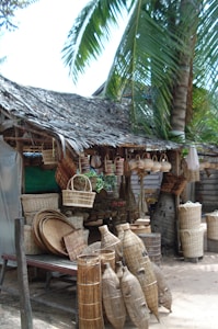 A rustic outdoor market stall displays a variety of woven baskets and fish traps, all made from natural materials. The setting includes a thatched roof made from palm leaves, providing shade. A large palm tree adds to the tropical atmosphere, and several different styles of baskets and traps are neatly arranged on tables.
