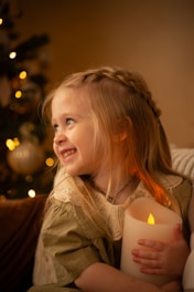 A young girl with braided blonde hair is smiling brightly while holding a large glowing candle. She is wearing a dress with a lace collar, and the background features a Christmas tree adorned with lights and ornaments. The scene is warmly lit, creating a cozy and festive atmosphere.