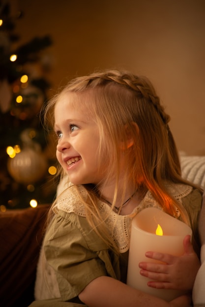 A young girl with braided blonde hair is smiling brightly while holding a large glowing candle. She is wearing a dress with a lace collar, and the background features a Christmas tree adorned with lights and ornaments. The scene is warmly lit, creating a cozy and festive atmosphere.
