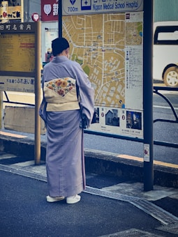 A person wearing traditional Japanese clothing, likely a kimono, stands in front of a large map display on a city street. The map provides directions to Nippon Medical School Hospital and other nearby locations. The individual is positioned with their back to the camera, facing the map. The clothing features a floral design on the back. Nearby, there is vehicular traffic and several signs written in Japanese.