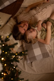 A woman and a child are lying on a cozy quilt with checkered pillows. They appear to be smiling and enjoying a moment together, adjacent to a Christmas tree adorned with lights.