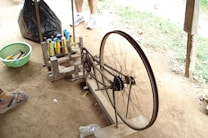 A spinning wheel setup is placed on a dirt floor with spools of thread in varied colors beside it. Several spools are arranged neatly on a rack near the wheel. In the background, there is a black plastic bag and a pair of white sneakers visible, likely worn by someone standing nearby.