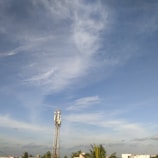 A tall communication tower stands against a backdrop of a clear blue sky with scattered white clouds. Several green palm trees and a few rooftops are visible in the foreground.