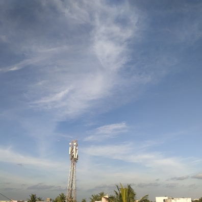 A tall communication tower stands against a backdrop of a clear blue sky with scattered white clouds. Several green palm trees and a few rooftops are visible in the foreground.