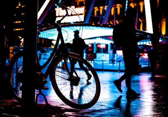 A silhouette of a bicycle is prominently displayed against a brightly lit, colorful urban background. Neon lights in shades of blue and red reflect off the wet pavement, creating a moody and vibrant atmosphere. Several people are walking in the background, their figures also cast in silhouette, adding to the dynamic city scene.