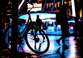 A silhouette of a bicycle is prominently displayed against a brightly lit, colorful urban background. Neon lights in shades of blue and red reflect off the wet pavement, creating a moody and vibrant atmosphere. Several people are walking in the background, their figures also cast in silhouette, adding to the dynamic city scene.