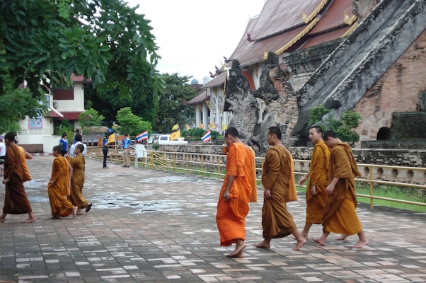 A group of monks dressed in orange robes walk along a paved area with a historic temple in the background. The temple features ornate architecture with dragon-like sculptures and traditional roofing. Flags line a railing alongside the temple, and lush greenery surrounds the area. People are moving in different directions, contributing to a sense of daily life at a religious site.