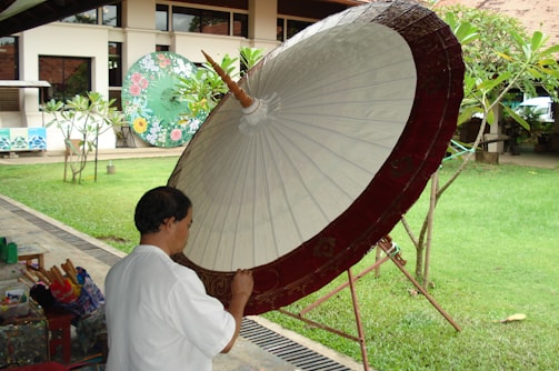 A person is painting a large, traditional paper umbrella with a red border, set outdoors with a background of green grass and plants. Additional colorful umbrellas are visible in the background, adding a decorative touch to the calm setting.