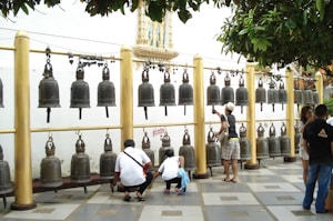 A row of large metallic bells hanging from a horizontal beam supported by golden pillars forms the focal point. Several people, including a child, are interacting with the bells, either examining them closely or taking photos. The setting appears to be outdoor, with leafy branches partially framing the image from above.