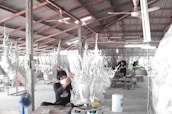 A workshop environment with individuals working on elaborate, artistic sculptures. The sculptures have intricate, flowing designs and the setting appears utilitarian with a corrugated metal roof and a concrete floor. The workers are focused and wearing protective masks.