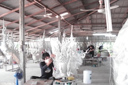 A workshop environment with individuals working on elaborate, artistic sculptures. The sculptures have intricate, flowing designs and the setting appears utilitarian with a corrugated metal roof and a concrete floor. The workers are focused and wearing protective masks.