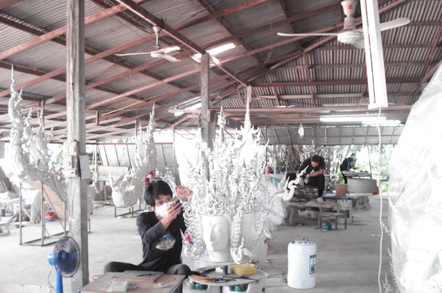 A workshop environment with individuals working on elaborate, artistic sculptures. The sculptures have intricate, flowing designs and the setting appears utilitarian with a corrugated metal roof and a concrete floor. The workers are focused and wearing protective masks.