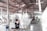 A workshop environment with individuals working on elaborate, artistic sculptures. The sculptures have intricate, flowing designs and the setting appears utilitarian with a corrugated metal roof and a concrete floor. The workers are focused and wearing protective masks.