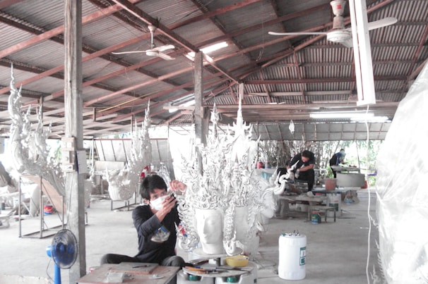 A workshop environment with individuals working on elaborate, artistic sculptures. The sculptures have intricate, flowing designs and the setting appears utilitarian with a corrugated metal roof and a concrete floor. The workers are focused and wearing protective masks.