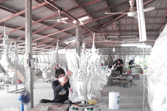 A workshop environment with individuals working on elaborate, artistic sculptures. The sculptures have intricate, flowing designs and the setting appears utilitarian with a corrugated metal roof and a concrete floor. The workers are focused and wearing protective masks.