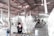 A workshop environment with individuals working on elaborate, artistic sculptures. The sculptures have intricate, flowing designs and the setting appears utilitarian with a corrugated metal roof and a concrete floor. The workers are focused and wearing protective masks.