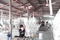 A workshop environment with individuals working on elaborate, artistic sculptures. The sculptures have intricate, flowing designs and the setting appears utilitarian with a corrugated metal roof and a concrete floor. The workers are focused and wearing protective masks.