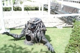A detailed statue of a creature with a reptilian head and elaborate armor emerging from the ground, set on a grassy area. The background features benches and a paved walkway in a garden-like setting.