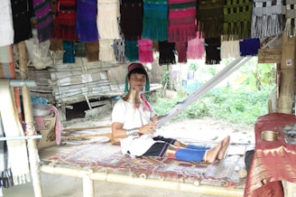 A person wearing traditional attire with multiple neck rings is seated on a bamboo platform, weaving fabric. Colorful textiles hang overhead, and the setting appears to be outdoors with greenery visible in the background.