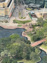 Aerial view of an urban area that includes a large building complex, pathways, roads, and a partially visible water feature surrounded by trees. A construction site with machinery and a crane can also be seen, alongside a park area with lush greenery.
