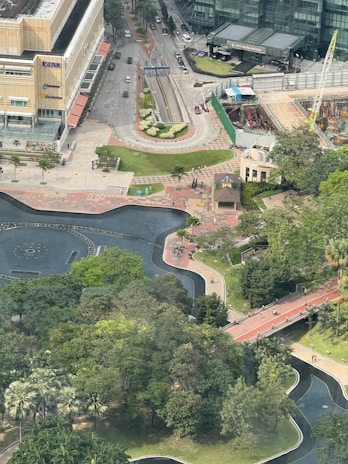 Aerial view of an urban area that includes a large building complex, pathways, roads, and a partially visible water feature surrounded by trees. A construction site with machinery and a crane can also be seen, alongside a park area with lush greenery.