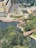 Aerial view of an urban area that includes a large building complex, pathways, roads, and a partially visible water feature surrounded by trees. A construction site with machinery and a crane can also be seen, alongside a park area with lush greenery.