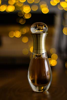 A perfume bottle with an elegant, elongated shape sits on a surface. The bottle is topped with a spherical lid and has a golden, ribbed neck. In the background, circular bokeh lights in warm yellow hues create an artistic and sophisticated ambiance.