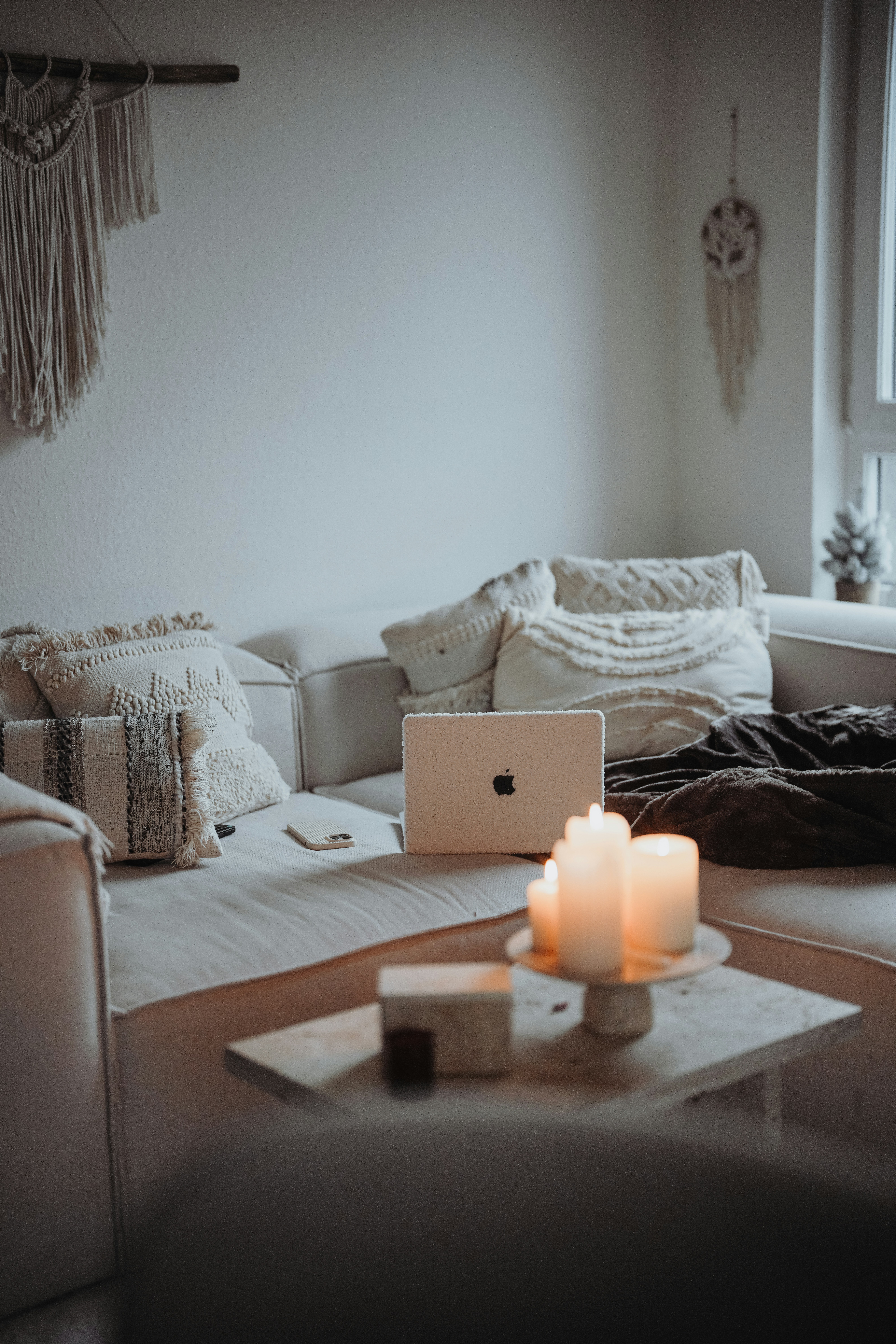 A cozy and minimalist living room area featuring a white sofa with textured cushions. A laptop is placed on the sofa, surrounded by a closed notebook and a smartphone. On the small coffee table in front, several lit candles create a warm and inviting atmosphere. Wall decor includes a hanging macrame piece and a dreamcatcher.