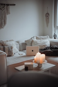 A cozy and minimalist living room area featuring a white sofa with textured cushions. A laptop is placed on the sofa, surrounded by a closed notebook and a smartphone. On the small coffee table in front, several lit candles create a warm and inviting atmosphere. Wall decor includes a hanging macrame piece and a dreamcatcher.