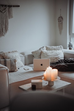 A cozy and minimalist living room area featuring a white sofa with textured cushions. A laptop is placed on the sofa, surrounded by a closed notebook and a smartphone. On the small coffee table in front, several lit candles create a warm and inviting atmosphere. Wall decor includes a hanging macrame piece and a dreamcatcher.