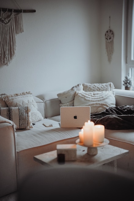 A cozy and minimalist living room area featuring a white sofa with textured cushions. A laptop is placed on the sofa, surrounded by a closed notebook and a smartphone. On the small coffee table in front, several lit candles create a warm and inviting atmosphere. Wall decor includes a hanging macrame piece and a dreamcatcher.