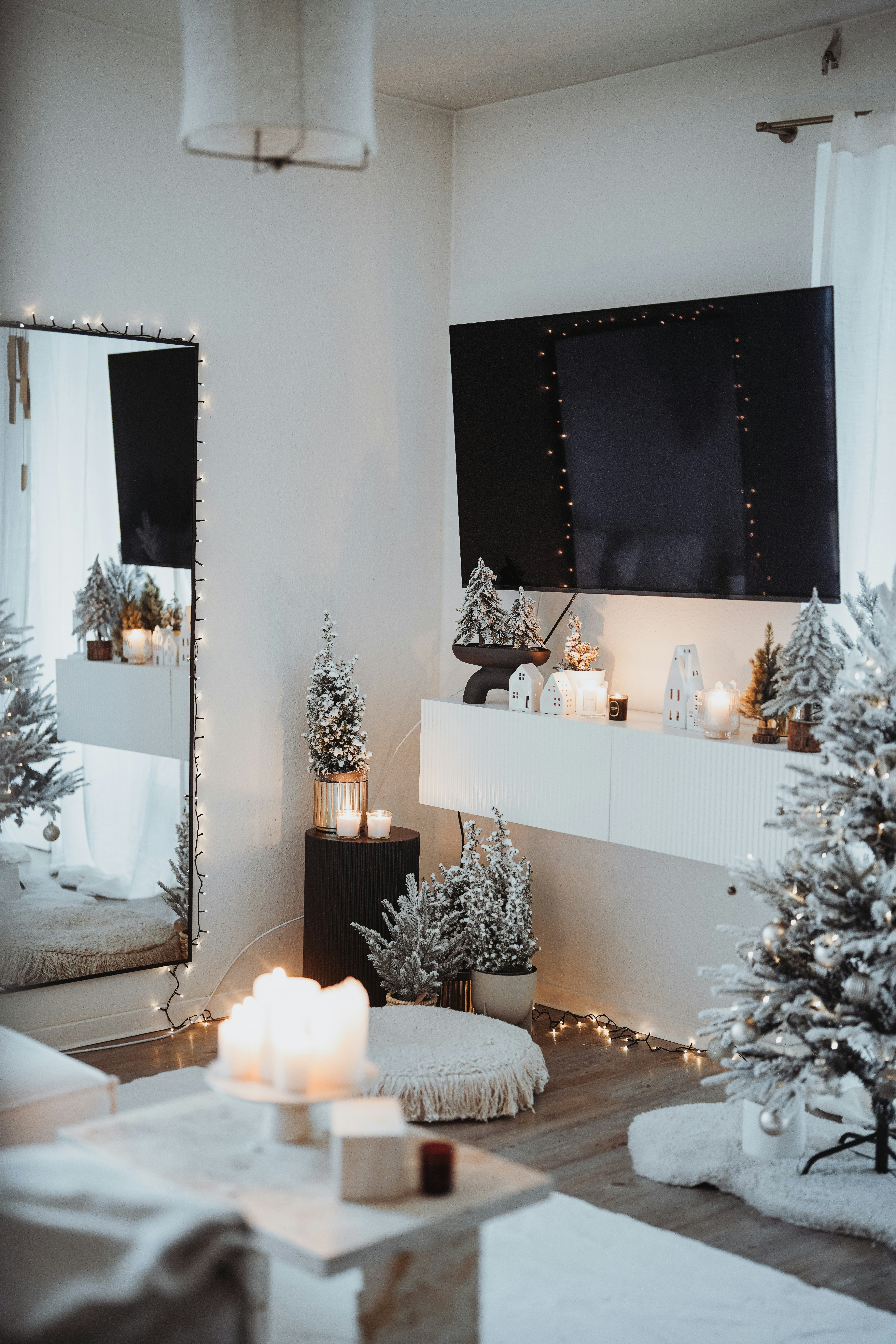 A cozy, elegantly decorated room with a winter or holiday theme. A large wall-mounted TV is adorned with string lights. Below it, a shelf holds small decorative houses with lights inside. Snow-dusted artificial trees, a plush rug, and a pouf contribute to the warm atmosphere. There are lit candles on a table and on the ledge beneath the TV. The overall ambiance is inviting and festive.