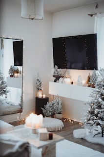 A cozy, elegantly decorated room with a winter or holiday theme. A large wall-mounted TV is adorned with string lights. Below it, a shelf holds small decorative houses with lights inside. Snow-dusted artificial trees, a plush rug, and a pouf contribute to the warm atmosphere. There are lit candles on a table and on the ledge beneath the TV. The overall ambiance is inviting and festive.