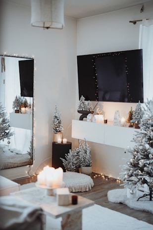 A cozy, elegantly decorated room with a winter or holiday theme. A large wall-mounted TV is adorned with string lights. Below it, a shelf holds small decorative houses with lights inside. Snow-dusted artificial trees, a plush rug, and a pouf contribute to the warm atmosphere. There are lit candles on a table and on the ledge beneath the TV. The overall ambiance is inviting and festive.