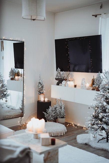A cozy, elegantly decorated room with a winter or holiday theme. A large wall-mounted TV is adorned with string lights. Below it, a shelf holds small decorative houses with lights inside. Snow-dusted artificial trees, a plush rug, and a pouf contribute to the warm atmosphere. There are lit candles on a table and on the ledge beneath the TV. The overall ambiance is inviting and festive.