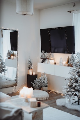 A cozy, elegantly decorated room with a winter or holiday theme. A large wall-mounted TV is adorned with string lights. Below it, a shelf holds small decorative houses with lights inside. Snow-dusted artificial trees, a plush rug, and a pouf contribute to the warm atmosphere. There are lit candles on a table and on the ledge beneath the TV. The overall ambiance is inviting and festive.