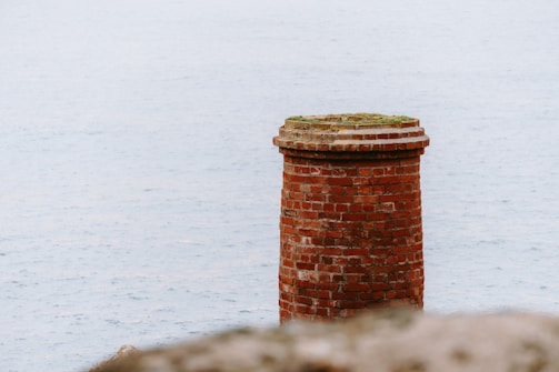 A solitary red brick chimney stands against a backdrop of calm, expansive water. The chimney is cylindrical and weathered, with a slightly overgrown top, suggesting age and abandonment.