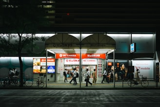 A nighttime city scene featuring a Bic Camera store with bright signage. Several people walk by under the illuminated storefront. The scene includes bicycles parked along the sidewalk, with a tree on the left side adding greenery to the urban setting.