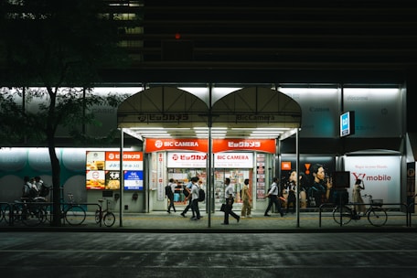 A nighttime city scene featuring a Bic Camera store with bright signage. Several people walk by under the illuminated storefront. The scene includes bicycles parked along the sidewalk, with a tree on the left side adding greenery to the urban setting.