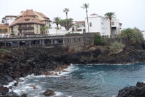 The image depicts a coastal scene with rocky cliffs and a small waterfront area. Several buildings with red-tiled roofs and white walls are situated on top of the cliffs, surrounded by palm trees and other greenery. The sea is a deep blue color with white waves crashing onto the rocks.