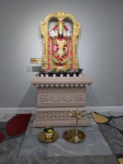 A richly decorated golden statue stands atop an intricately carved pedestal. Surrounding the statue are vibrant garlands of flowers in red and green. The pedestal features ornamental designs and symbols. In front of the pedestal: a brass oil lamp and a small container placed on a tray, enhancing the spiritual atmosphere.