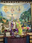 A vibrant and detailed depiction of deities wearing ornate, colorful garments and adornments, set against a backdrop featuring deities in the sky, animals like cows, deer, and peacocks, as well as lush greenery. Floral arrangements and offerings are placed in the foreground.