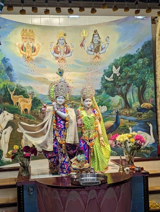 A vibrant and detailed depiction of deities wearing ornate, colorful garments and adornments, set against a backdrop featuring deities in the sky, animals like cows, deer, and peacocks, as well as lush greenery. Floral arrangements and offerings are placed in the foreground.