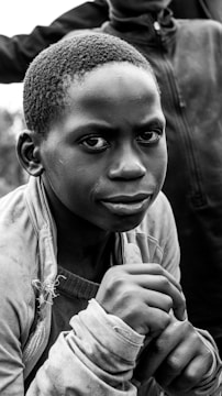 A close-up of a young person looking directly at the camera with an introspective expression. The black and white image captures detailed textures, including a worn jacket and a thoughtful look in the person's eyes.