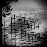 A silhouetted steel framework of a building under construction is set against a dramatic sky filled with textured clouds. Sunlight peeks through the gaps, creating a stark contrast with the dark structure. Silhouetted leaves and branches from nearby trees border the scene.